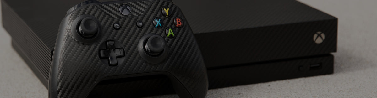 A close-up of a black Xbox One S console with a carbon-fiber textured skin on the controller, set against a dark background.