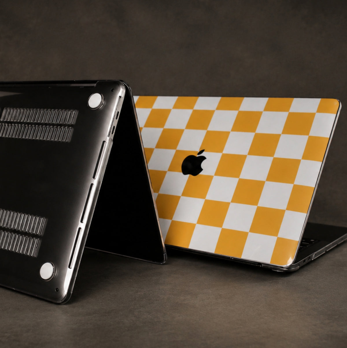 Two Apple MacBook laptops shown on a dark studio surface, one displaying the black underside and the other showing a yellow and white checkerboard pattern skin on the back of the lid.