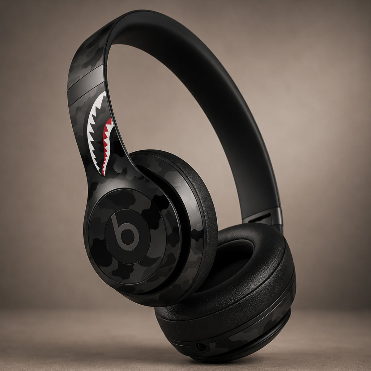Black Beats Studio3 wireless headphones with a dark camouflage pattern and a shark mouth graphic on the headband, shown on a neutral brown background.