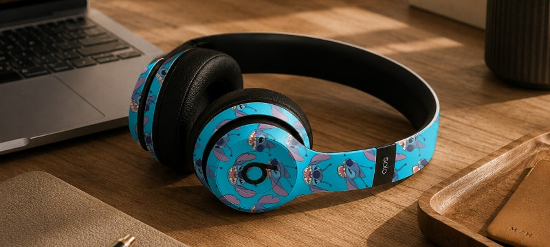 Blue over-ear headphones with a cartoon character pattern, resting on a wooden desk next to a laptop.