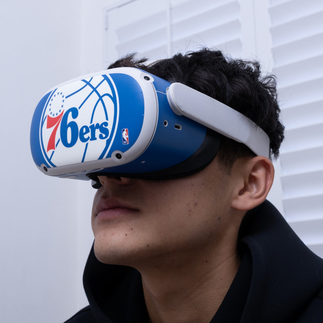 A man wearing a blue and white Philadelphia 76ers-branded VR headset, shown in profile against a white window background.