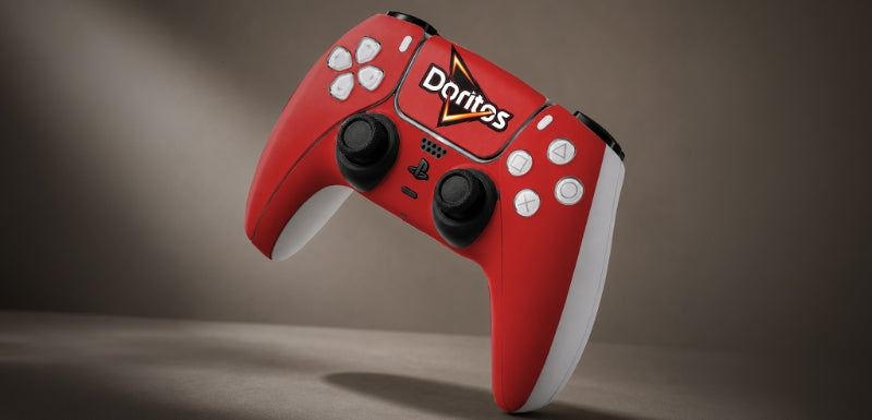 A red PlayStation-style game controller with white grips and black joysticks, featuring the Doritos logo on the top center, shown against a dark brown studio background.