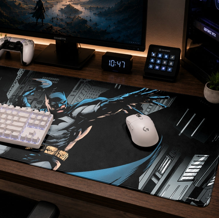 A large Batman-themed mousepad on a wooden desk, styled with a keyboard, mouse, digital clock, and monitor in a gaming setup.