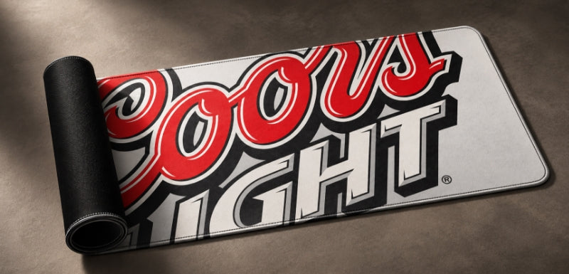 A Coors Light branded rectangular mat or rug shown partially unrolled on a dark brown textured surface. The visible top side features the large red, white, and black Coors Light logo, and the rolled edge reveals a black underside.